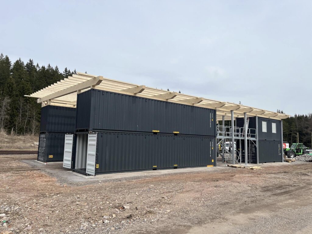 Das Foto zeigt den Rohbau unserer Container-Werkstatt von schräg vorne. Auf einer hellgrauen Schotterfläche stehen acht dunkelgraue See-Container, welche teilweise von Gitterlaufstegen durchbrochen werden. Jeweils vier Container stehen auf je einer Seite des Reparaturgleises. Auf dem Foto ist gut zu sehen, dass die Container durch große Türen an ihren Stirnseiten begehbar sind.