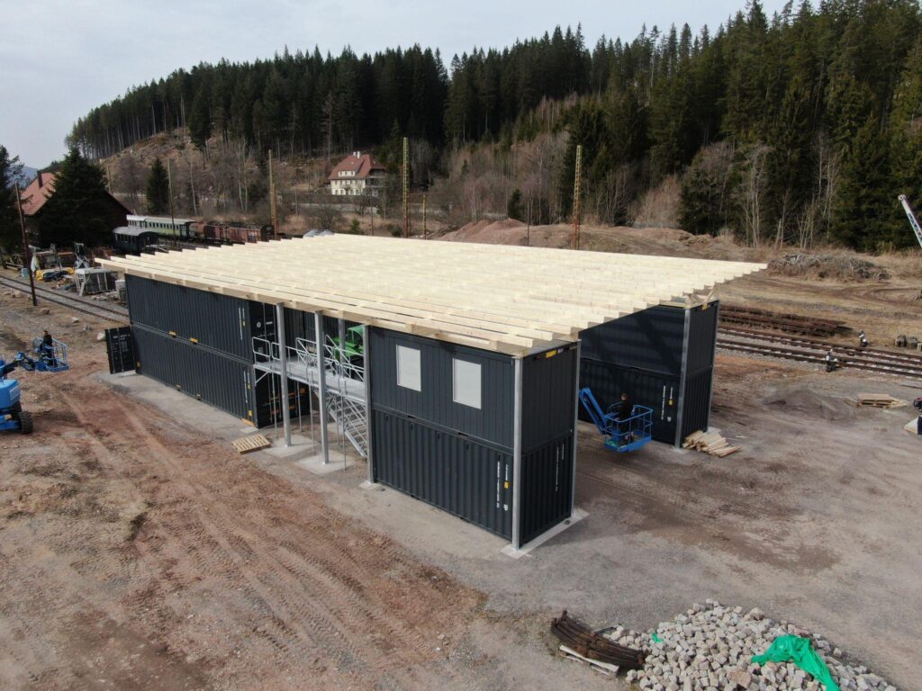 Das Foto zeigt den Rohbau unserer Container-Werkstatt von schräg oben. Auf einer hellgrauen Schotterfläche stehen acht dunkelgraue See-Container, welche teilweise von Gitterlaufstegen durchbrochen werden. Jeweils vier Container stehen auf je einer Seite des Reparaturgleises, die gesamte Konstruktion wird von einem Schrägdach aus Holz überspannt.