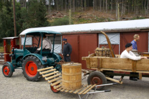 Das Bild wurde während des Bahnhofsfests aufgenommen. Auf dem Bild ist links ein grüner Hanomag-Traktor zu sehen, der vor einem gedeckten Güterwaggon steht. Der Traktor zieht einen Holzanhänger, auf dem ein Strohballen steht und eine Person sitzt. Im Vordergrund steht eine alte Schubkarre, die mit einem Holzfass beladen ist.