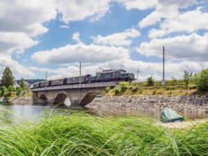 Auf dem Bild zieht eine alte E94 Elektro-Lokomotive einen Personenzug von links nach rechts über die Eisenbahnbrücke am Schluchsee. Die Brücke überspannt mit drei niedrigen Bögen einen Teil des Schluchsees.