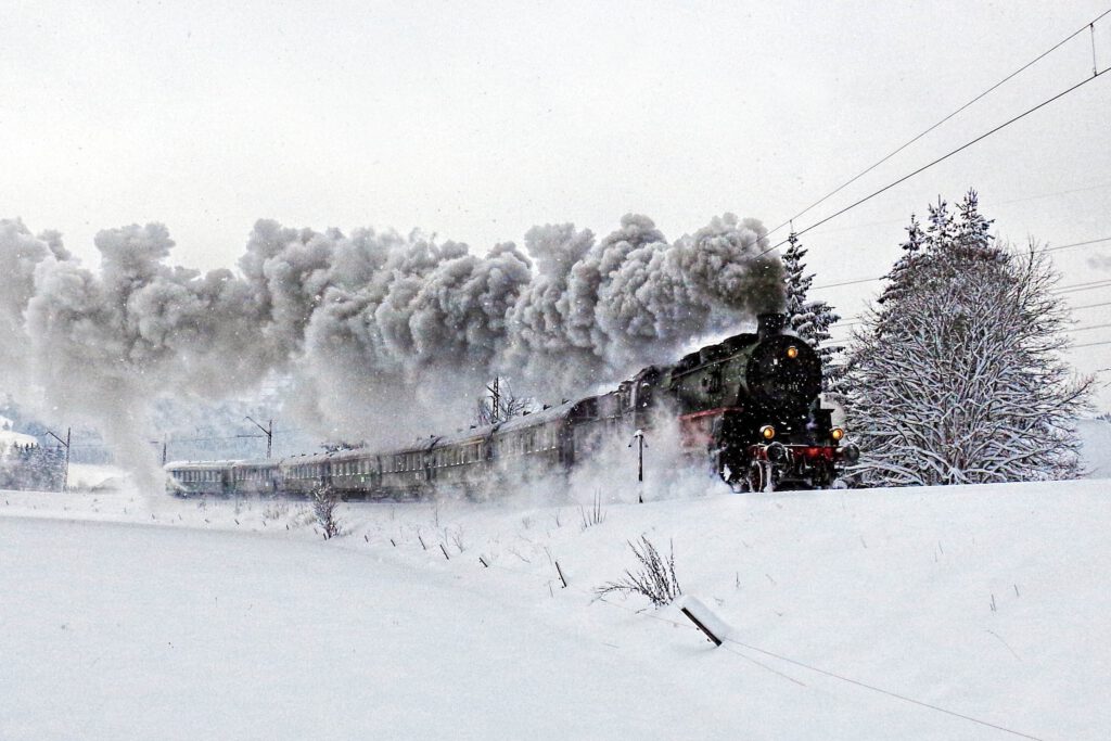 Auf dem Bild ist eine verschneite Landschaft zu sehen. Auf einem ebenfalls verschneiten Bahndamm kommt von links nach rechts eine Dampflokomotive angefahren, die mehrere grüne Personenwaggons sieht.