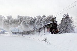 Auf dem Bild ist eine verschneite Landschaft zu sehen. Auf einem ebenfalls verschneiten Bahndamm kommt von links nach rechts eine Dampflokomotive angefahren, die mehrere grüne Personenwaggons sieht.