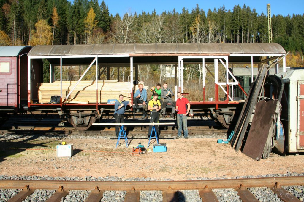 Eine Gruppe Freiwilliger der Drei-Seen-Bahn sitzt im Rohhbau eines Güterwaggons. Man sieht nur die Metallstreben der Seitenwände, die Holzverkleidung fehlt. Im Vordergrund liegen Werkzeuge und Materialien.