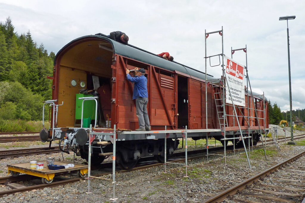 Auf dem Foto sieht man einen gedeckten Güterwaggon, dessen Seitentüren geöffnet sind. Sowohl auf dem Dach als auch an der Seite sind Vereinsmitglieder der Drei-Seen-Bahn damit beschäftigt, Reparaturen auszuführen.