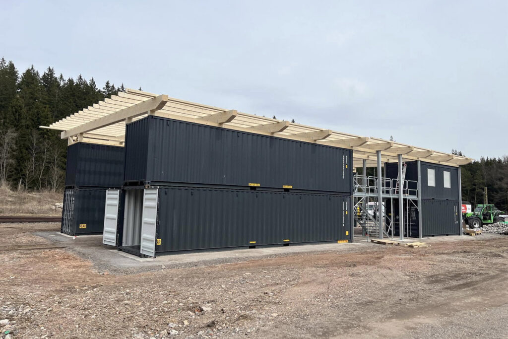 Das Foto zeigt den Rohbau unserer Container-Werkstatt von schräg vorne. Auf einer hellgrauen Schotterfläche stehen acht dunkelgraue See-Container, welche teilweise von Gitterlaufstegen durchbrochen werden. Jeweils vier Container stehen auf je einer Seite des Reparaturgleises. Auf dem Foto ist gut zu sehen, dass die Container durch große Türen an ihren Stirnseiten begehbar sind.
