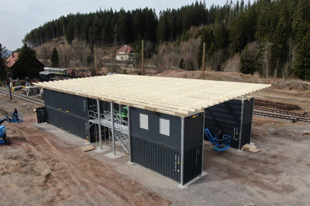 Das Foto zeigt den Rohbau unserer Container-Werkstatt von schräg oben. Auf einer hellgrauen Schotterfläche stehen acht dunkelgraue See-Container, welche teilweise von Gitterlaufstegen durchbrochen werden. Jeweils vier Container stehen auf je einer Seite des Reparaturgleises, die gesamte Konstruktion wird von einem Schrägdach aus Holz überspannt.